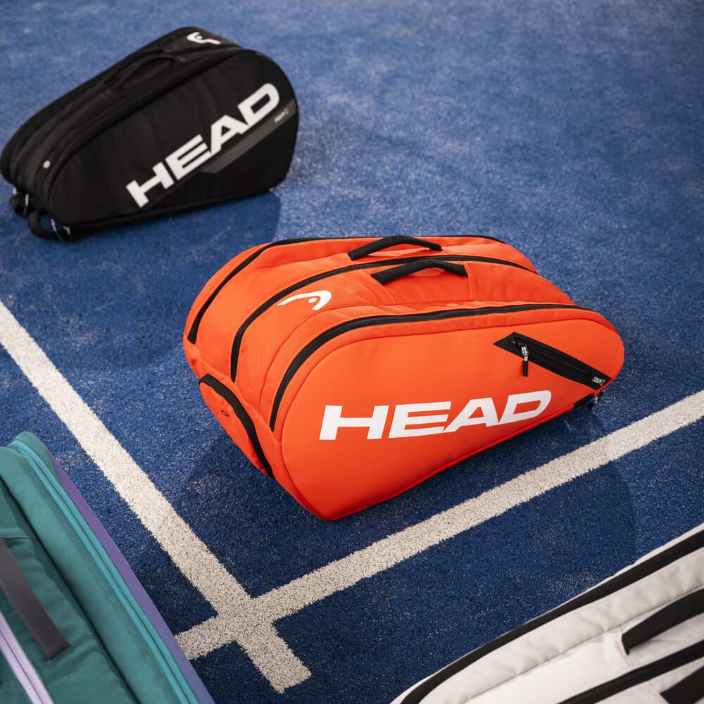 A luminous fluo orange HEAD Tour Padel Bag L is positioned on a blue tennis court amidst other sports bags. It boasts a backpack carry system and CCT+ climate control to ensure your gear remains in excellent condition.