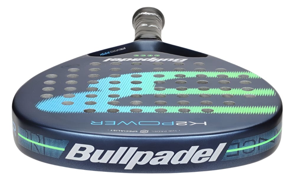 Close-up of a Bullpadel K2 Power 2025 padel racket against a gray background. The blue racquet, with a carbon fiber frame, features a circular head with black and turquoise accents, perforations, and the brand name elegantly displayed in white on the frame and handle for power and control.