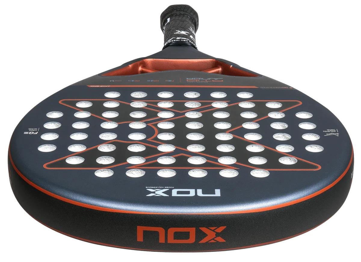 Close-up of a NOX AT10 Genius Junior By Augustin Tapia 2025 Padel Racket featuring a carbon frame, black and orange design with circular holes, textured grip handle, and the NOX logo on both the head and edge.