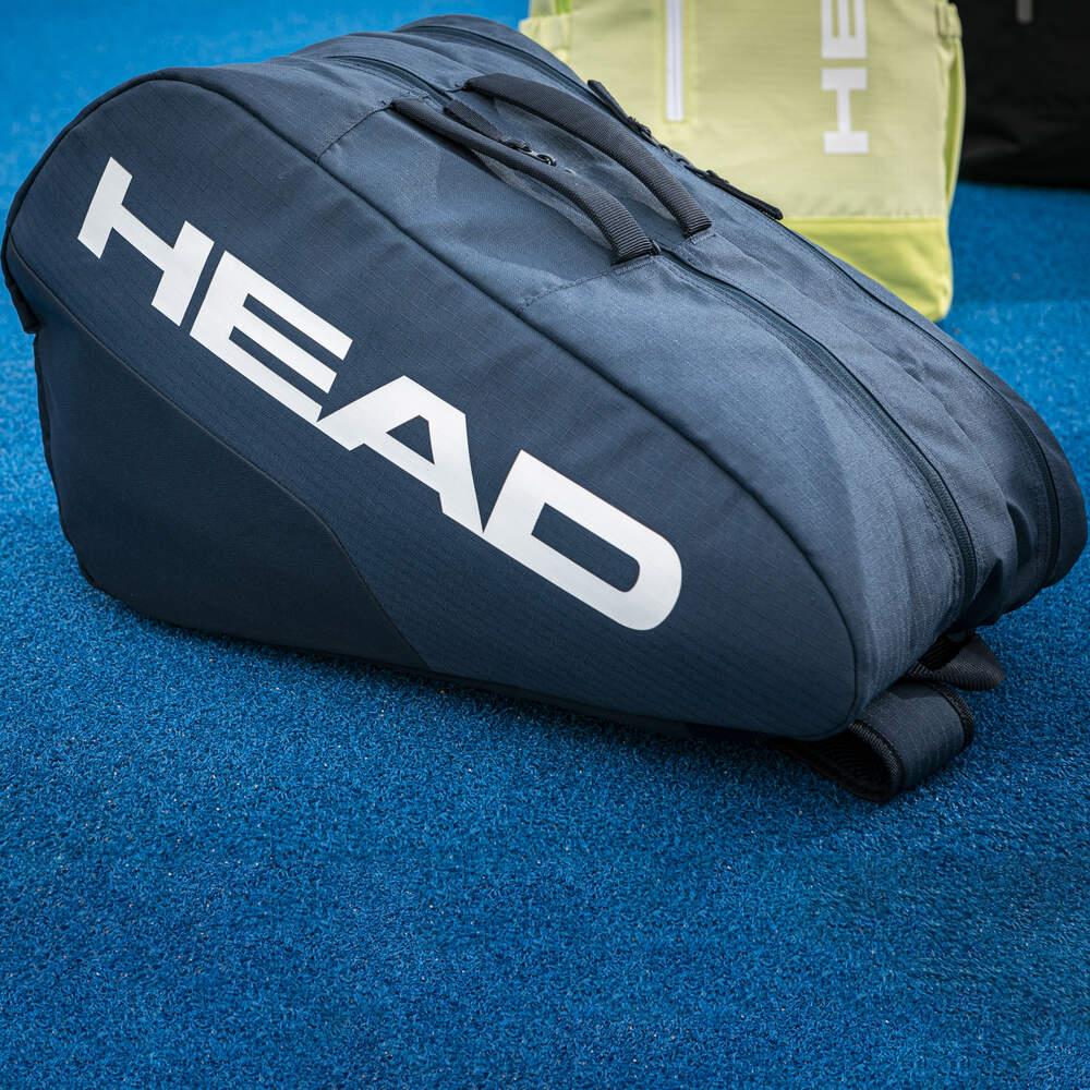 A HEAD Base Padel Racket Bag M in navy, featuring "HEAD" in bold white letters, rests on a blue surface. A light green bag is partially visible in the background. Both bags, designed for carrying sports equipment, feature reinforced construction.