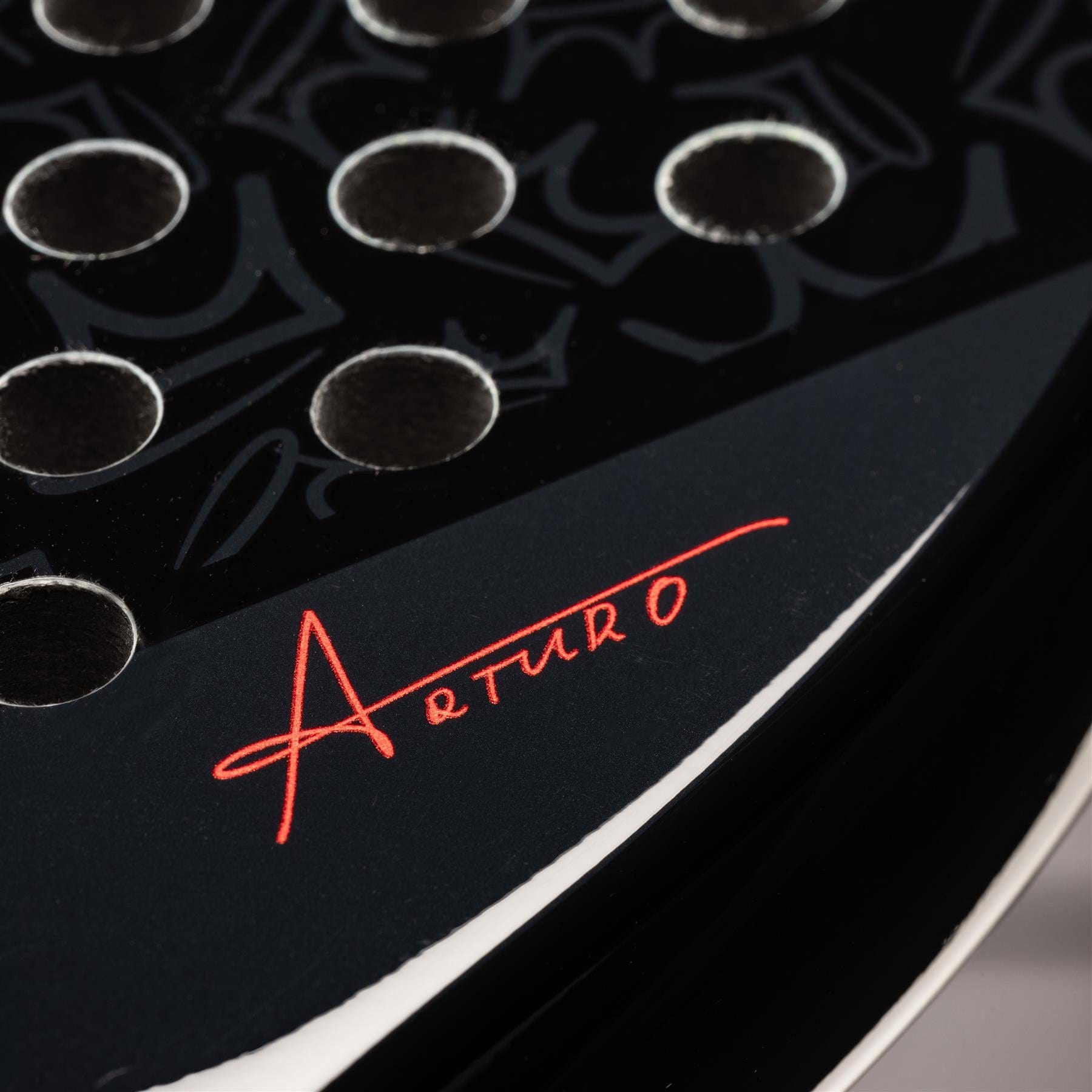 Close-up of the HEAD Coello Vibe 2025 Padel Racket in black, featuring round holes and a red Arturo Coello signature on its glossy surface. Subtle patterns in the background highlight this HEAD racket, ideal for beginners.