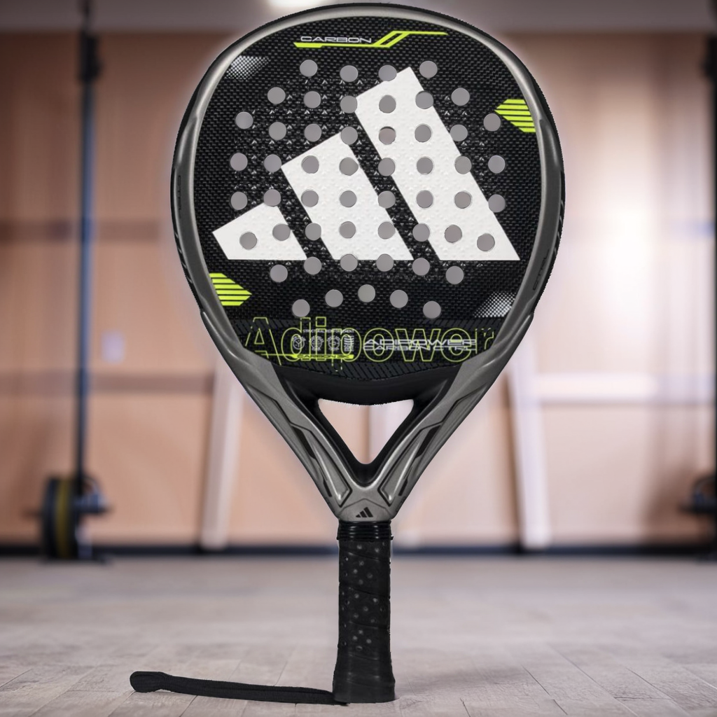 The adidas ADIDAS Adipower Carbon CTRL 3.4 Padel Racket in black and grey, featuring the signature "Adipower" logo and advanced technologies, stands upright on a gym floor with blurred fitness equipment in the background.