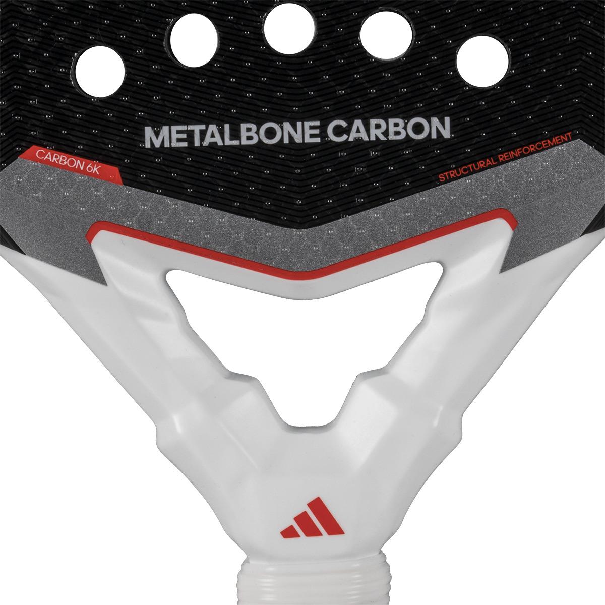 Close-up of the ADIDAS Metalbone Carbon 3.4 Padel Racket, ideal for advanced players. It has a black surface with circular holes and "METALBONE CARBON" text, while its textured white handle with red accents displays "CARBON 6K" and "STRUCTURAL REINFORCEMENT.