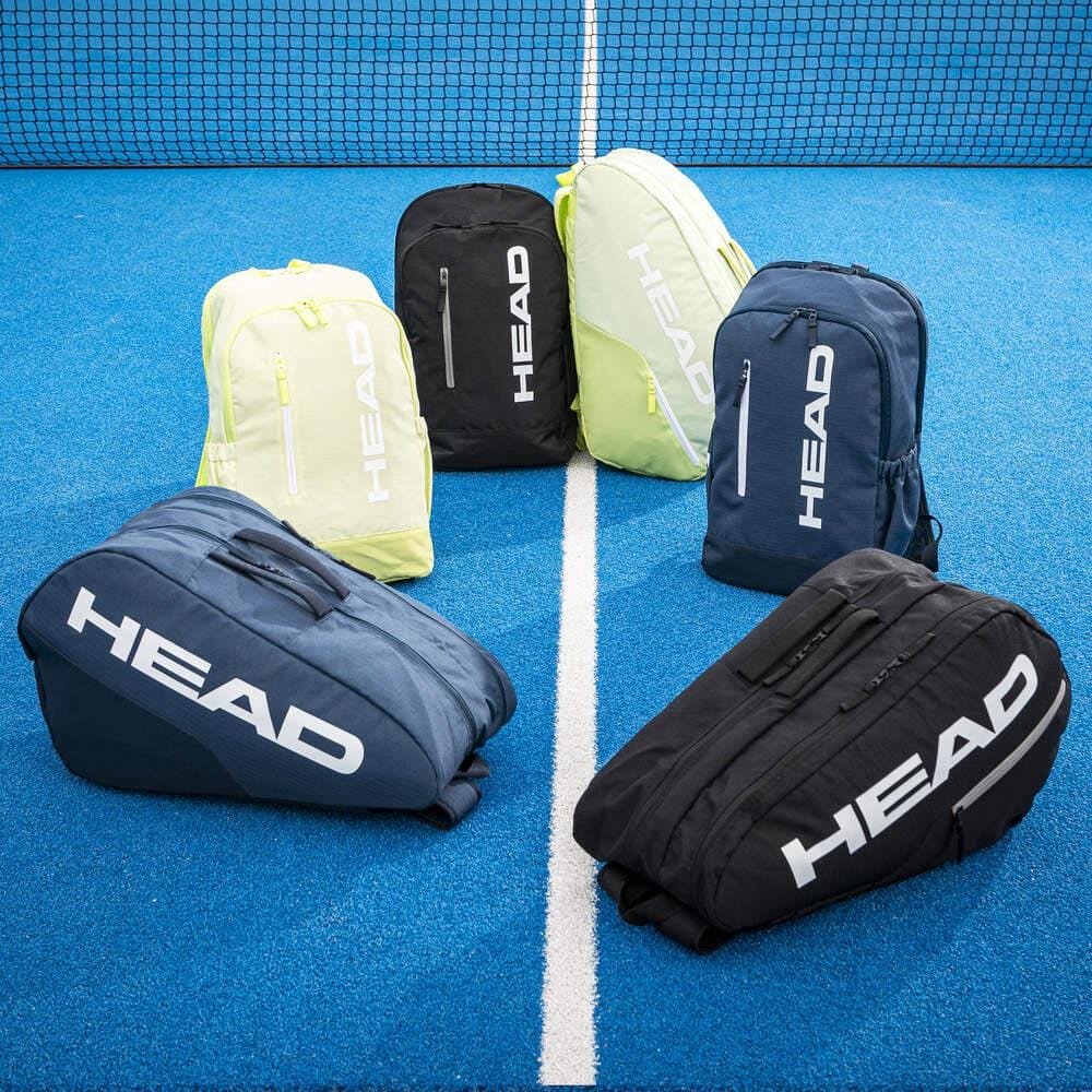 Six HEAD BASE Padel Backpack 17L bags in black are arranged in a semicircle on a bright blue tennis court, with the net visible in the background.