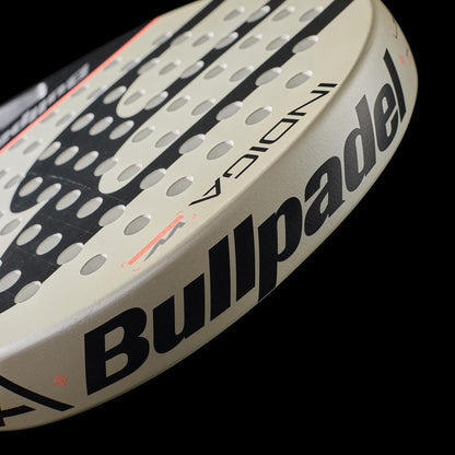 Close-up of the Bullpadel Indiga W 2026 Padel Racket, featuring lightweight performance, black "Bullpadel" and "INDIGA" text, and multiple round holes on its beige face, set against a black background.