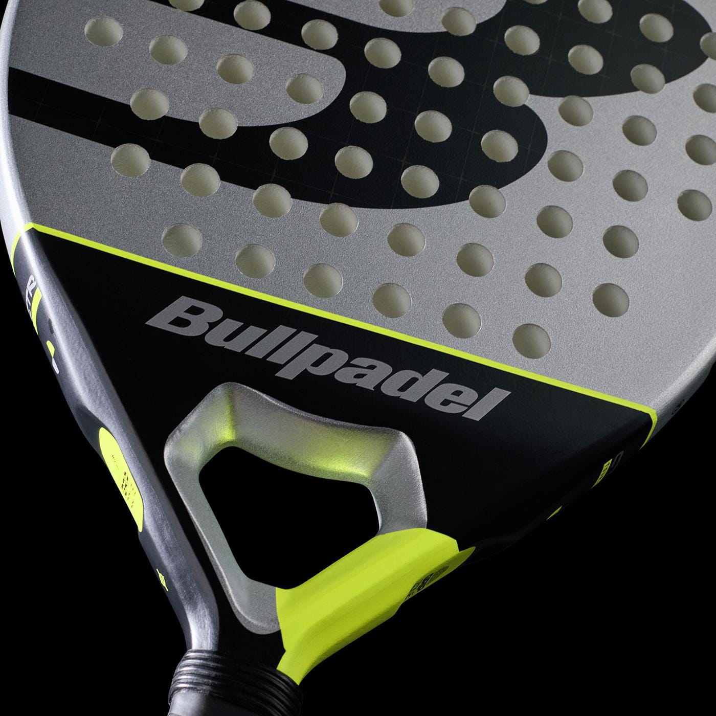 Close-up of the Bullpadel Indiga Ctr 2026 Padel Racket by Bullpadel, showcasing its silver and black face with round holes and yellow accents, crafted for precision control against a sleek black background.