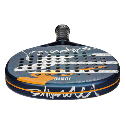 Close-up of the Bullpadel Ionic Control 25 Padel Racket, featuring a blue design with multiple holes, white abstract patterns, and signatures. Enhanced by the XForce system, it has an orange accent and black grip tape.