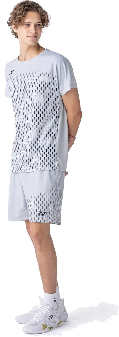 A young person with curly hair stands sideways, wearing a light gray sports t-shirt and Yonex 15241EX Knit Mens Padel Shorts in Silver Grey, paired with white athletic shoes.