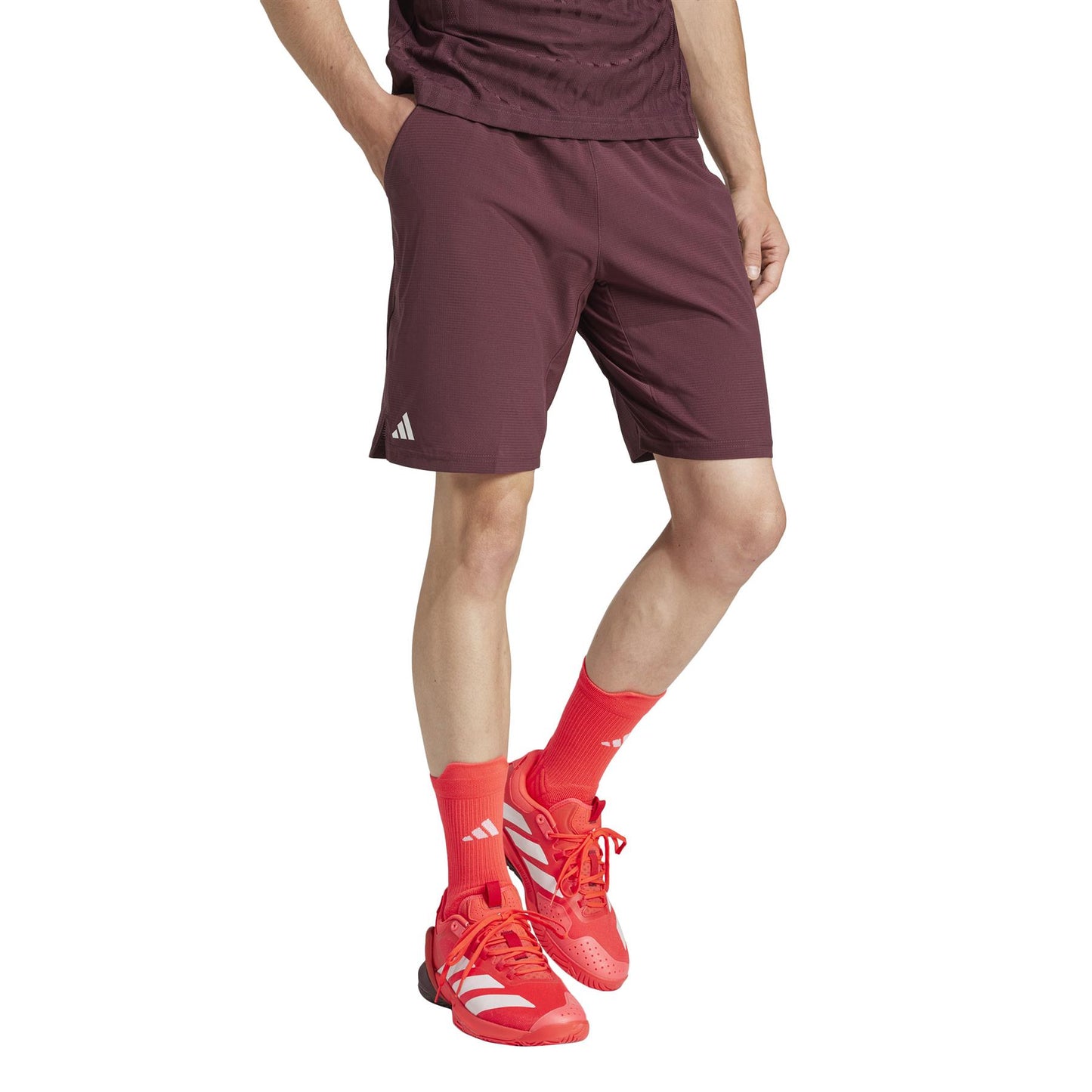 Dressed in ruby ADIDAS Ergo Men's Padel Shorts and a coordinated top, matched with vibrant red socks and red sneakers featuring white stripes, an individual poses against a plain white backdrop. The sporty, casual outfit is crafted from recycled materials.