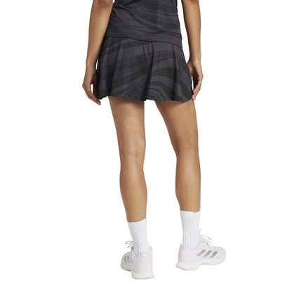 A person in a black Adidas Women's Club Graphic Padel Skirt, likely made from recycled materials, stands with their back to the camera. They wear white socks and sneakers, featuring Climacool technology, against a plain white background.