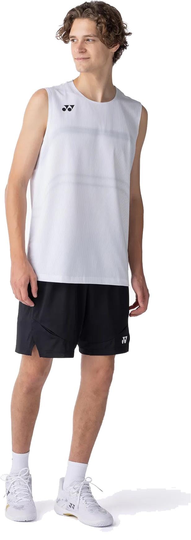 A young man with curly hair wears a sleeveless white athletic shirt and Yonex 15239YX Knit Mens Padel Shorts in black, paired with white sneakers. He stands slightly to the side, looking off-camera, in a relaxed pose.