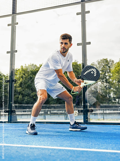 Dressed in white, a man gets ready to strike a padel ball with the HEAD Extreme Pro 2026 Padel Racket, highlighting its powerful design on an outdoor blue court bordered by glass walls and trees—an ideal choice from HEAD for advanced players who demand precision and strength.