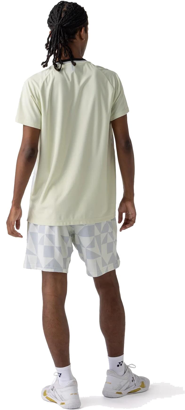 A person with braided hair faces away, wearing a light short-sleeve shirt, Yonex 15240YX Knit Mens Padel Shorts in Champagne, white socks, and white athletic shoes—all set against a white background.