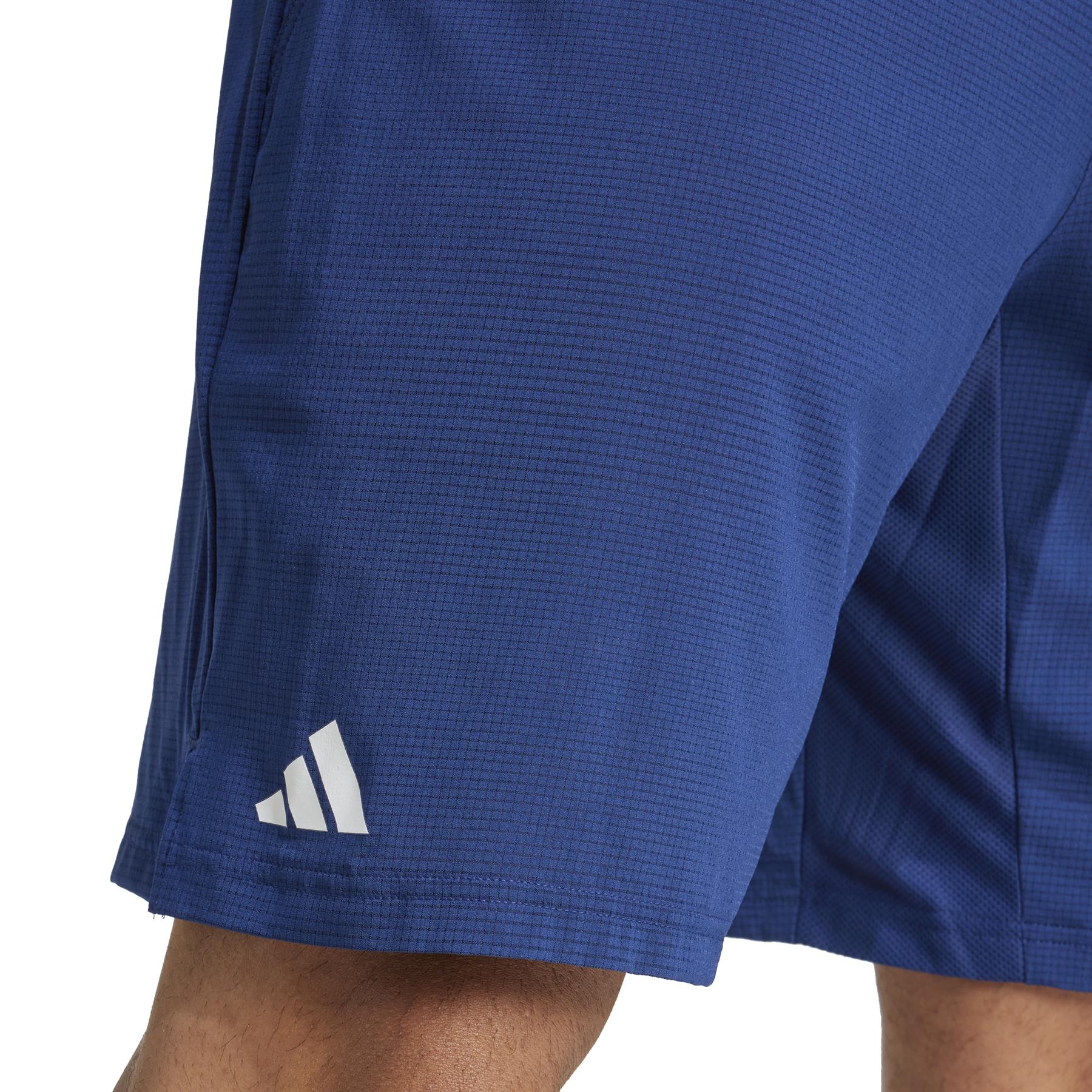 Someone wearing Adidas Ergo Men's Padel Shorts in navy with a white logo on the lower left. Made from recycled materials, the shorts have a textured pattern, and part of one leg is visible against a white background.