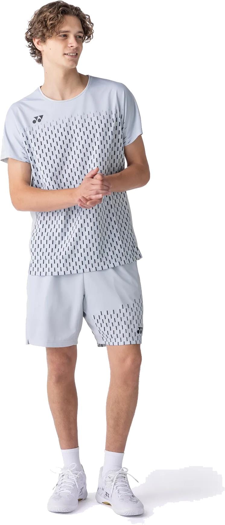 A young man faces left, wearing the Yonex 10713EX Crew Neck Men's Padel T-Shirt in Silver Grey with a black dashed pattern by Yonex, paired with white sneakers, gazing off to the side with a neutral expression.