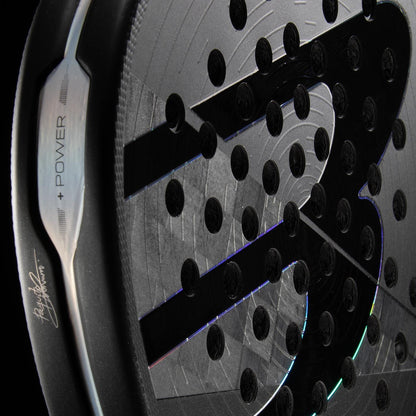 Close-up of the Bullpadel HACK 04 Hybrid 2026 Padel Racket in grey/black, featuring a textured surface, perforated holes, and a large stylized “S” logo with metallic edge engraved "POWER" and a signature, capturing its dynamic power.