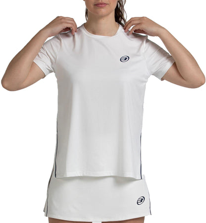 A woman wears the Bullpadel Picase Women's Padel T-Shirt in Bone, featuring moisture-wicking technology and small circular Bullpadel logos on the chest and skirt, adjusting her collar against a plain white background.
