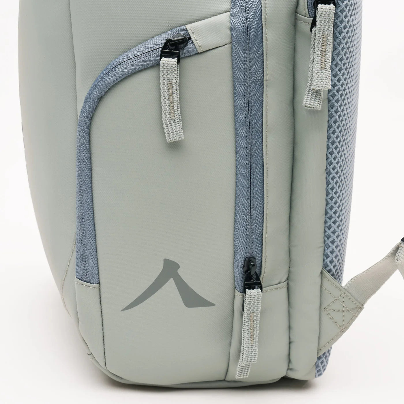 Close-up of a Kanso Intuko Padel 23L Backpack's side pocket in grey, featuring a zipper with fabric pull tab, textured section, and light blue accents. A subtle tilted triangle logo appears on the lower part alongside reflective print details.