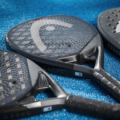 Three HEAD Speed One 2025 black padel rackets lie on a blue textured court. The image highlights the intricate design, logo patterns, and Auxetic 2.0 technology on their heads and handles.