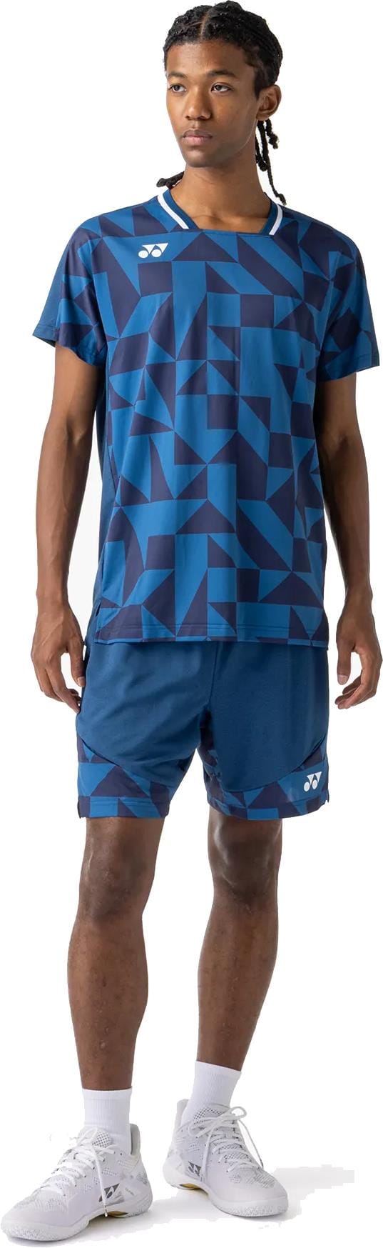 A person in a blue geometric sports shirt, Yonex 15240YX Knit Mens Padel Shorts - Indigo Blue, white crew socks, and white athletic shoes stands facing slightly to the side.