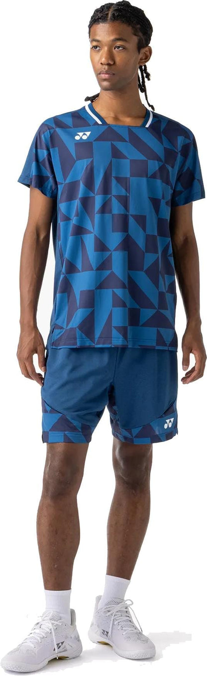 A person in a blue geometric sports shirt, Yonex 15240YX Knit Mens Padel Shorts - Indigo Blue, white crew socks, and white athletic shoes stands facing slightly to the side.