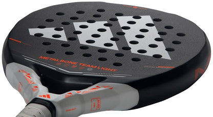 A close-up of the adidas Metalbone Team Light 2026 Padel Racket - Black for advanced players, showing a large white adidas logo, many circular holes, and "METALBONE TEAM LIGHT" printed in orange on the surface.