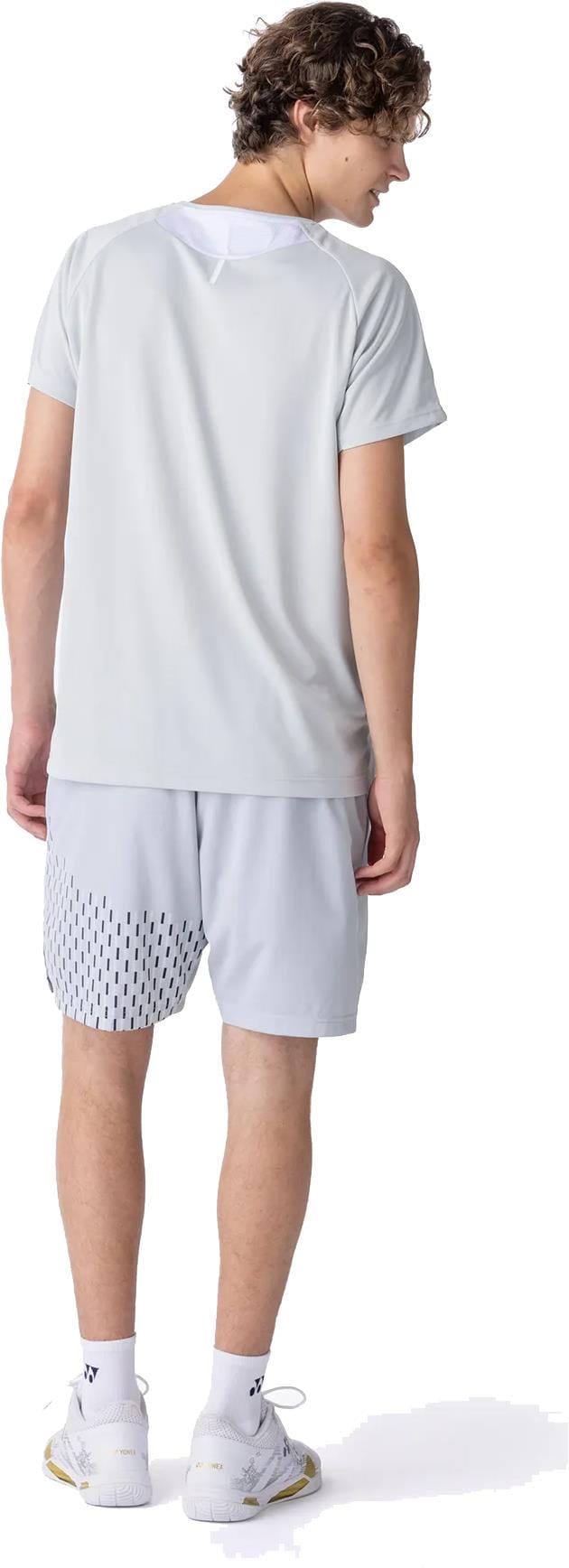 A young man with curly hair, turned away from the camera, wears a light gray athletic t-shirt and Yonex 15241EX Knit Mens Padel Shorts in Silver Grey, paired with white socks and sneakers.