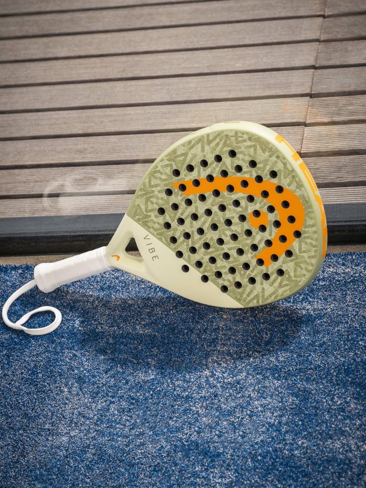 A HEAD Vibe 2026 Green / Orange Padel Racket, featuring a durable fibreglass hitting surface and "VIBE" on the handle, lies on a blue textured court near wood, with its orange logo displayed.