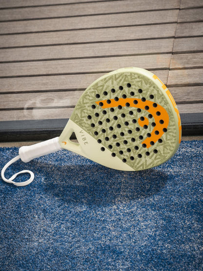 A HEAD Vibe 2026 Green / Orange Padel Racket, featuring a durable fibreglass hitting surface and "VIBE" on the handle, lies on a blue textured court near wood, with its orange logo displayed.