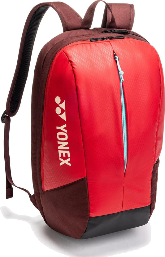 The Yonex 42512EX Team Padel Backpack in ruby red features a brown-red design, vertical blue front zipper, padded shoulder straps, black base, and white Yonex logo and name on the side. Ideal for racket sports.