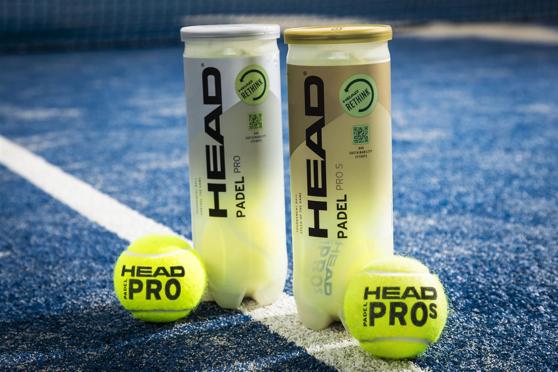 Two tubes of HEAD Padel Pro padel balls, each featuring lids in white and gold, rest on a blue court alongside two vibrant yellow padel balls. In the background is the silhouette of a net.