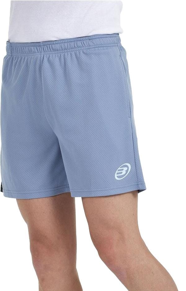 A person wearing Bullpadel Campaza Men's Padel Shorts in Shadow Blue, showing the white logo on the left leg and Quickerdry fabric, with an elastic waistband. Only their lower torso and upper legs are visible, paired with a white shirt.