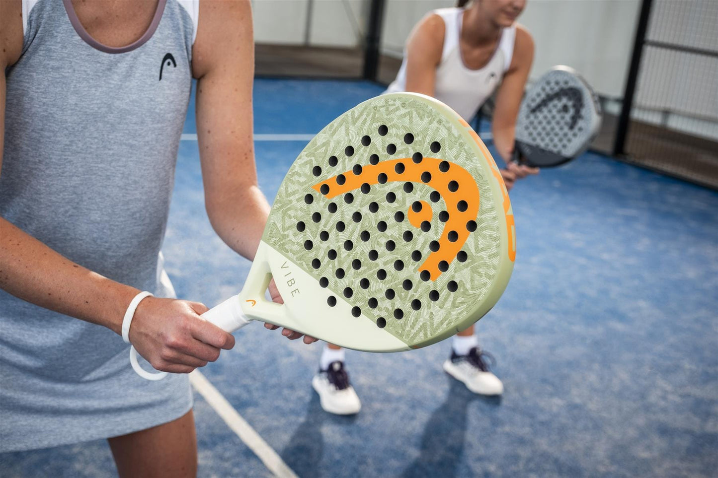 Two women in sportswear play padel on a blue court, highlighting the HEAD Vibe 2026 Green / Orange Padel Racket with fibreglass hitting surface in the foreground as they get ready to receive a serve, with another player and the net visible behind.