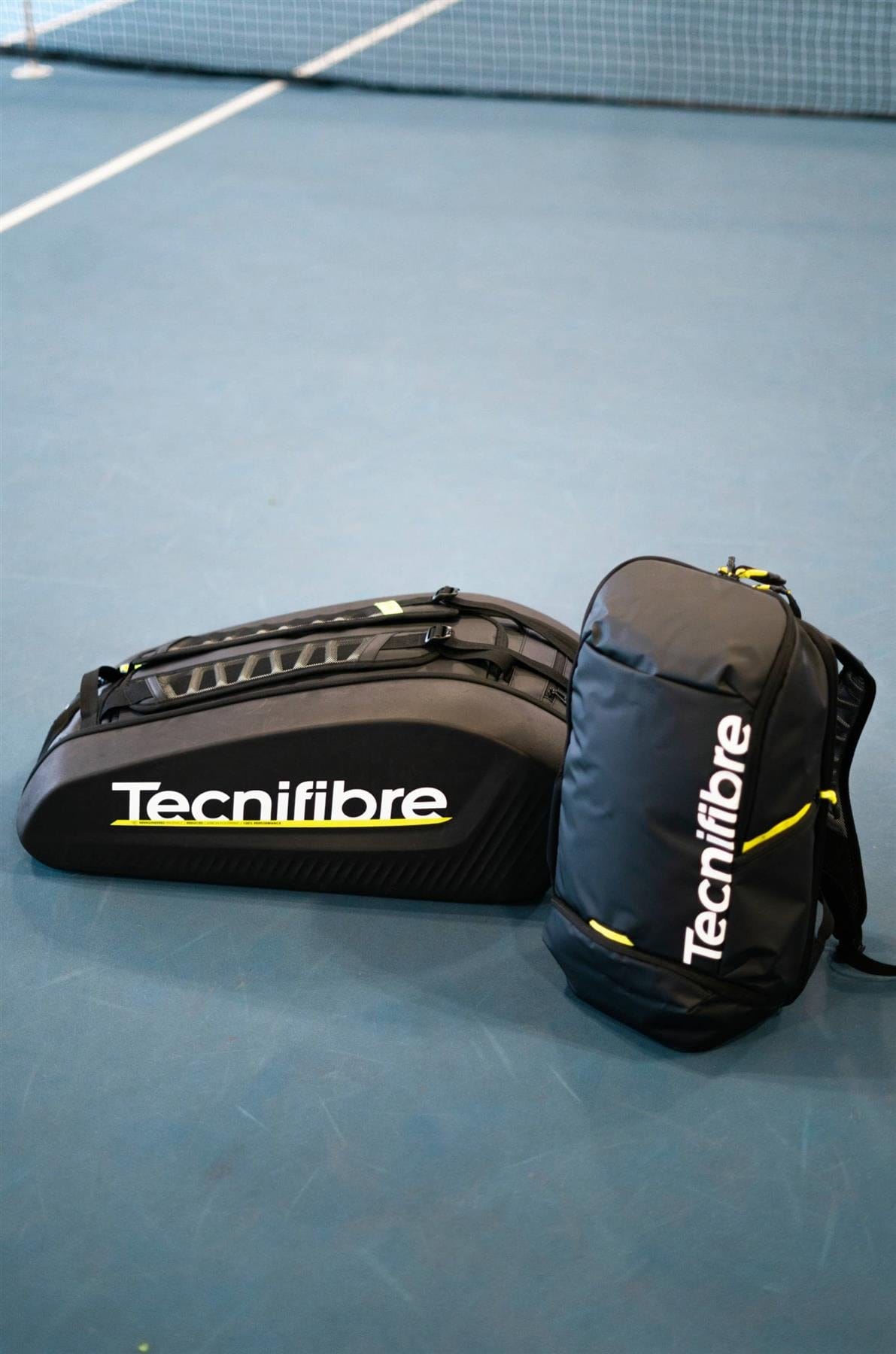 A black Tecnifibre Reform Padel Backpack and a Tecnifibre duffel bag sit on the blue tennis court near the net, both featuring yellow accents, white Tecnifibre logos, and durable EVA material for protection.