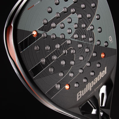 Close-up of the Bullpadel HACK 04 Comfort 2026 Padel Racket in grey, showcasing a textured surface with circular perforations and orange accents. The Bullpadel logo appears on the lower right side, reflecting its dynamic power and modern design.