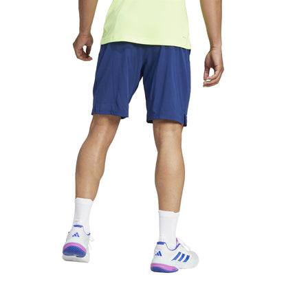 A person in a light green shirt and navy ADIDAS Ergo Men's Padel Shorts made from recycled materials stands against a white backdrop, wearing white socks and sneakers with purple and blue accents, seen from the back.