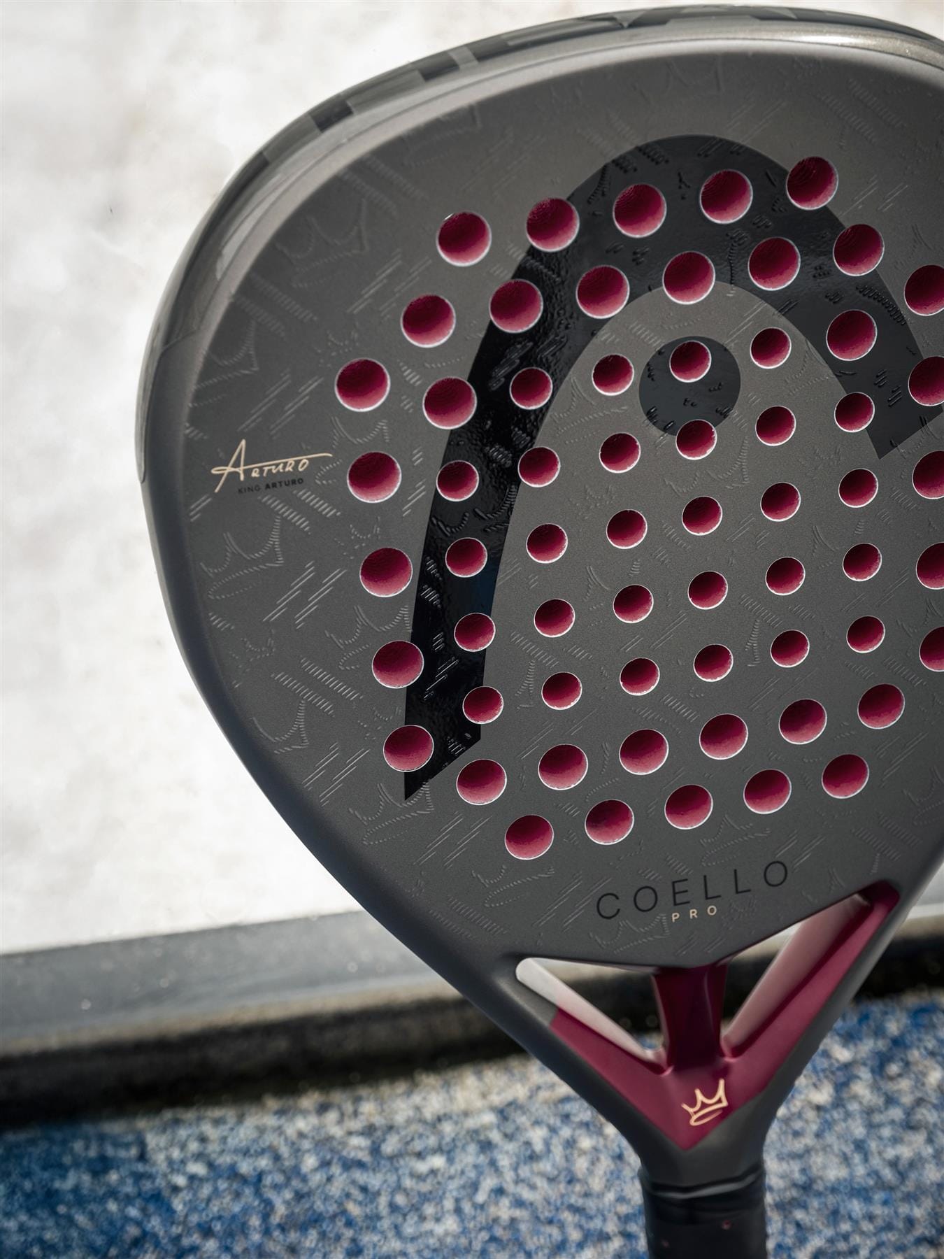 A close-up of the HEAD Coello Pro 2026 Padel Racket by HEAD, showcasing its carbon hybrid surface and striking black and magenta design, appears on a blue court with a blurred background.