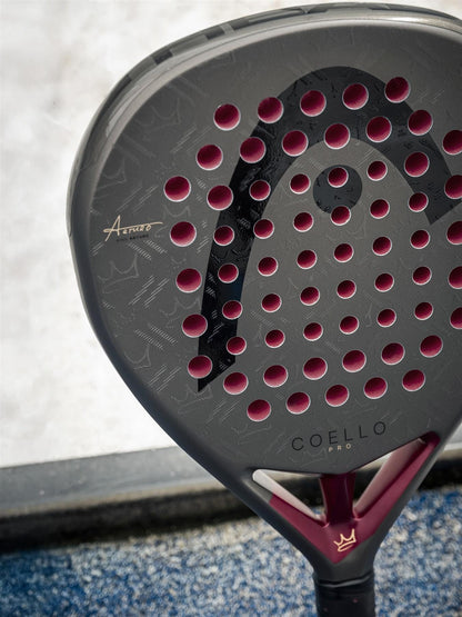 A close-up of the HEAD Coello Pro 2026 Padel Racket by HEAD, showcasing its carbon hybrid surface and striking black and magenta design, appears on a blue court with a blurred background.