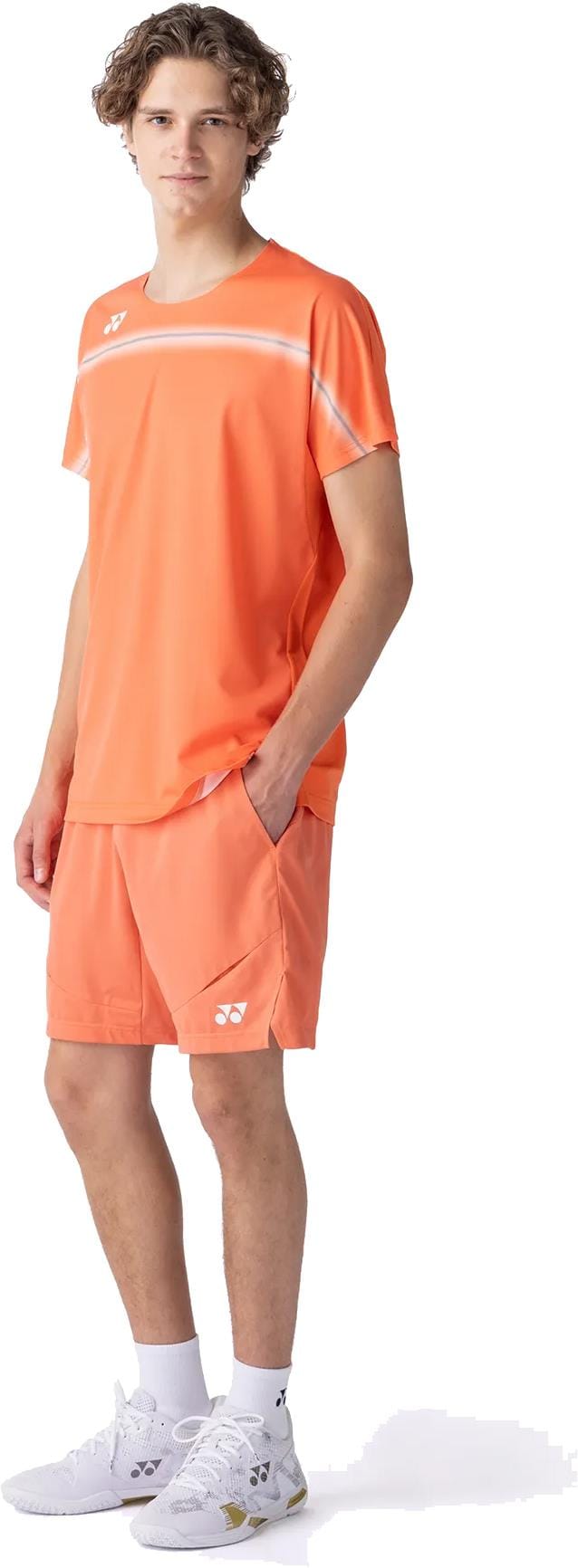 A young man with curly hair wears the Yonex 10711YX Crew Neck Mens Padel T-Shirt in Cyber Orange with matching shorts, white socks, and sneakers. He stands against a white background, one hand in his pocket, facing slightly to the side.