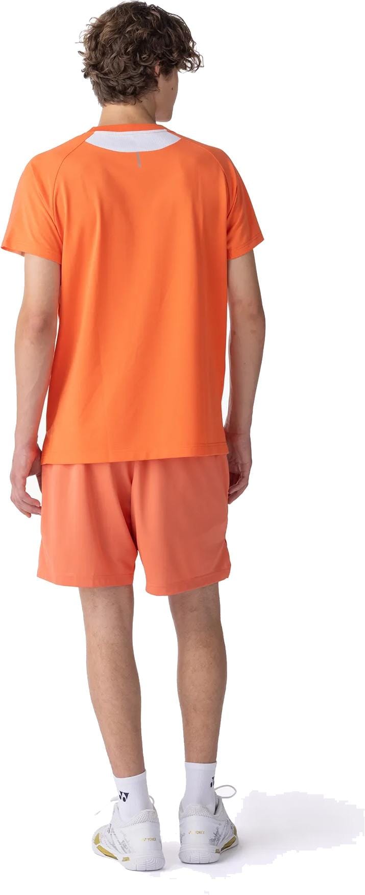 A young man with curly hair, wearing a Yonex 10711YX Crew Neck Mens Padel T-Shirt in Cyber Orange and matching shorts, stands with his back to the camera against a white background.