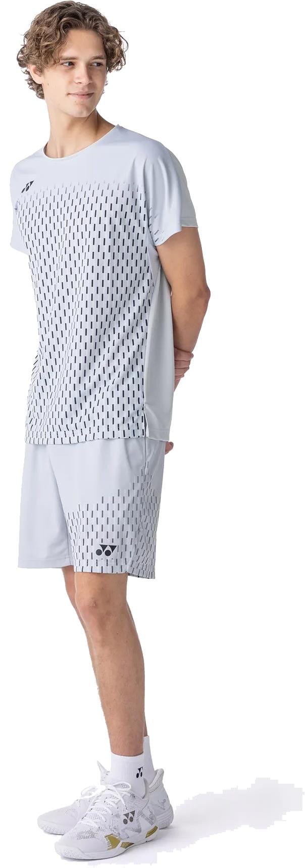 A young man with curly hair wears the Yonex 10713EX Crew Neck Mens Padel T-Shirt in Silver Grey, matching shorts, white socks, and white athletic shoes, standing with his hands behind his back and looking to the side.