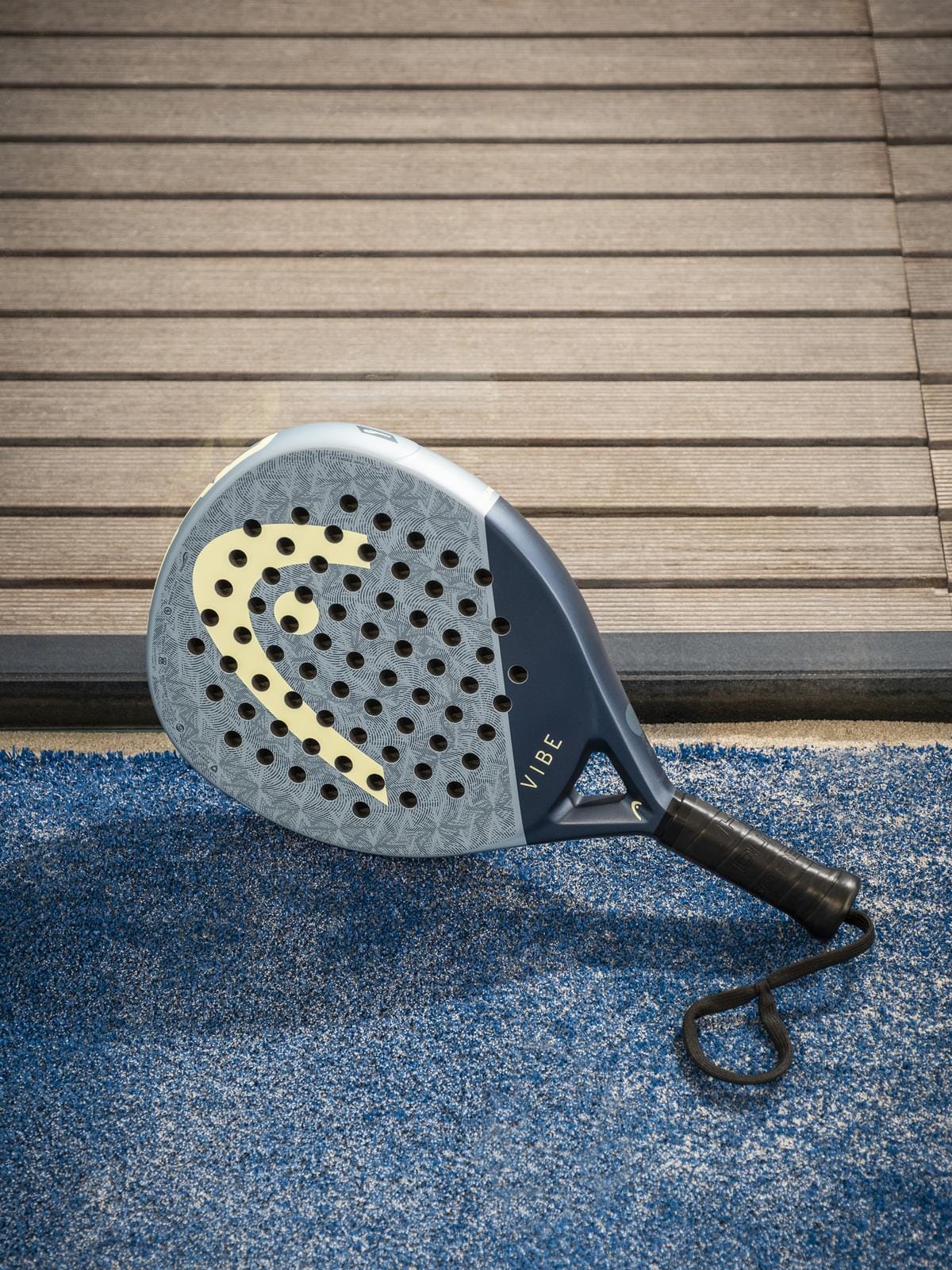 A HEAD Vibe 2026 Blue / Yellow Padel Racket by HEAD, with a fibreglass face and yellow logo, rests on blue artificial turf near wooden decking.