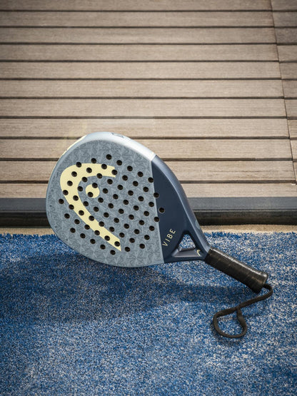 A HEAD Vibe 2026 Blue / Yellow Padel Racket by HEAD, with a fibreglass face and yellow logo, rests on blue artificial turf near wooden decking.