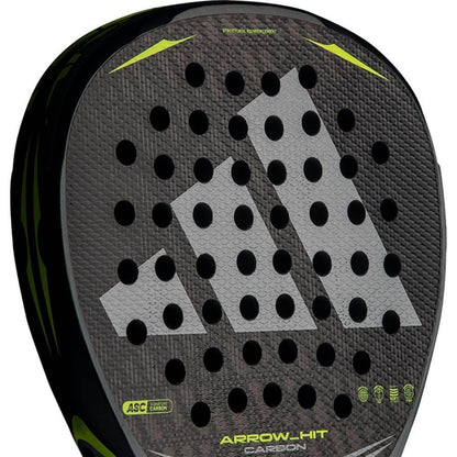 A close-up of the adidas Arrow Hit Carbon 2026 Padel Racket, showing its black and gray textured surface, round holes, neon yellow accents, and the adidas logo prominently displayed.
