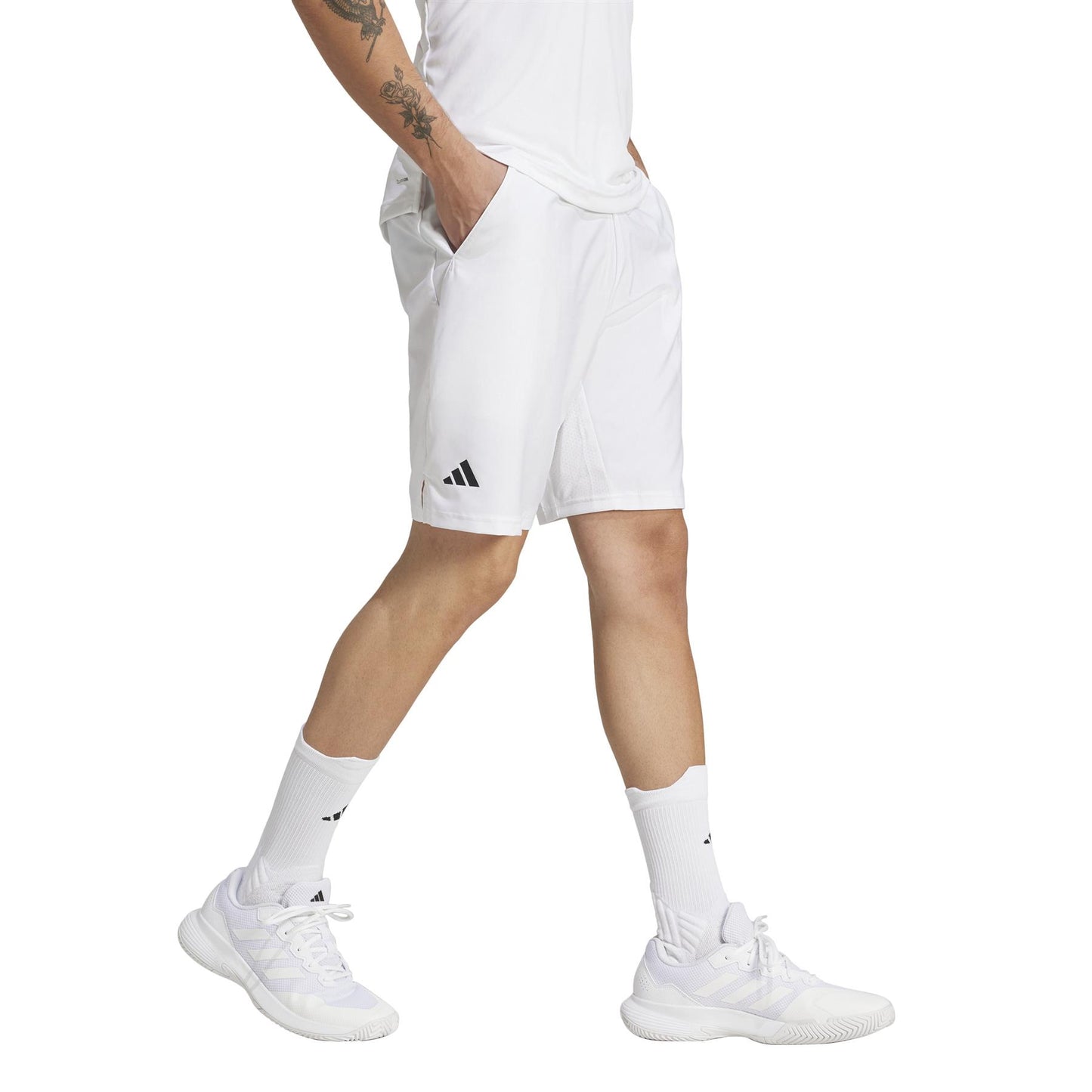 The person sports a white athletic outfit with a t-shirt, sneakers, and matching socks. The ADIDAS Men's 3 Stripe Padel Shorts feature a discreet logo near the hem and are made from recycled materials, ideal for an eco-conscious athlete with tattoos on their left forearm.