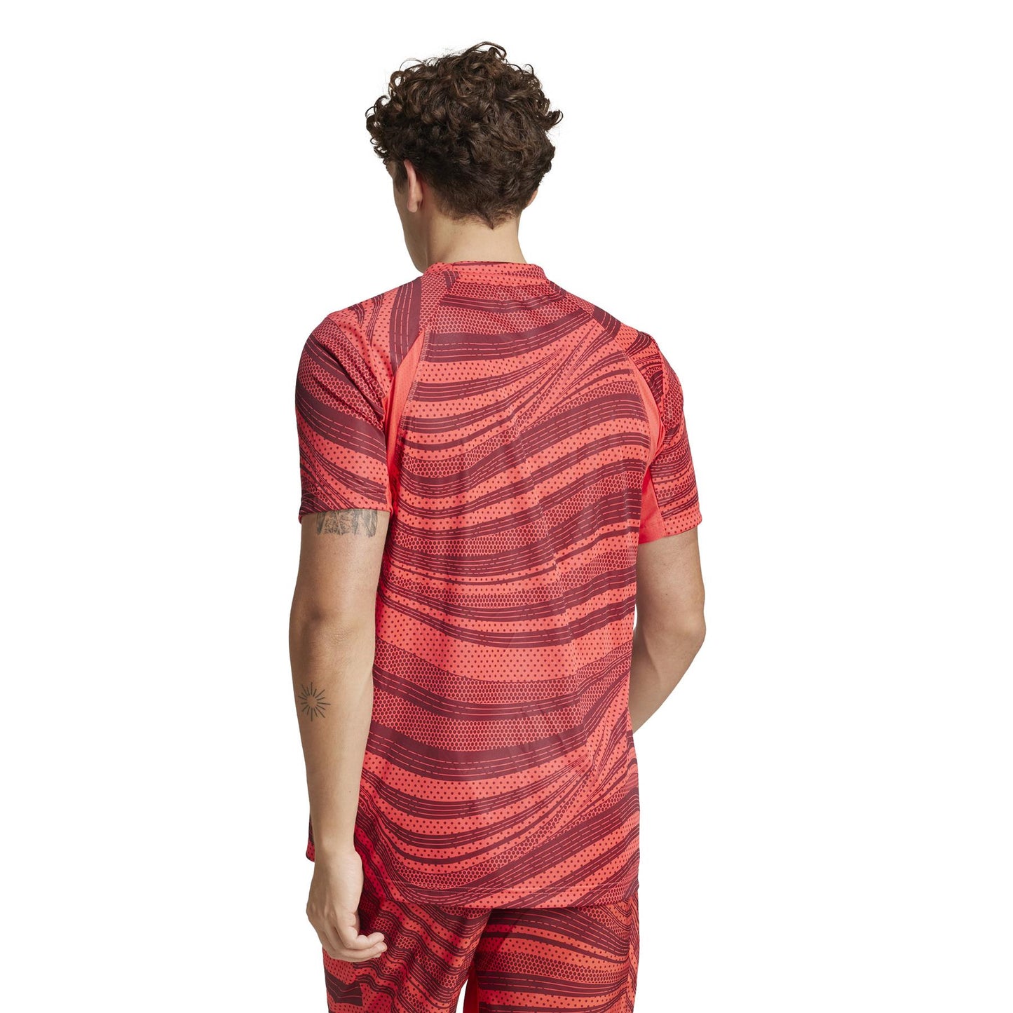 A curly-haired person, with their back to the camera, wears a red ADIDAS Men's Club Graphic Padel T-Shirt by adidas made from recycled materials. The dynamic red and black design pairs with matching shorts, and a small tattoo adorns their upper arm for a personalized flair.