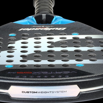 Close-up of the Bullpadel Vertex 05 Comfort 2026 Padel Racket by Bullpadel, featuring "CUSTOM WEIGHT SYSTEM" on its Fibrix fibre surface, circular holes, and a black-and-blue textured design.