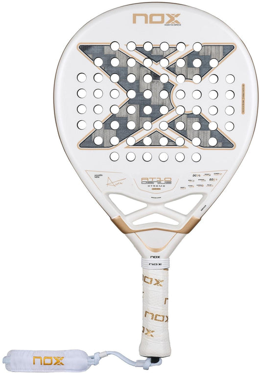 The NOX AT10 Genius 12K Alum Extrem By Agustín Tapia 2026 padel racket features a white frame with gold and gray accents, Tapia’s large "X" design, circular holes, and a white grip with "Nox" branding and matching wrist strap.