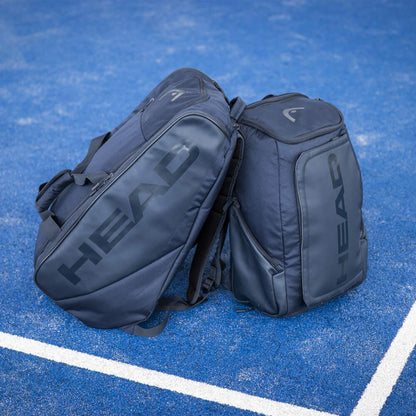 Two HEAD Pro Padel Bags L in navy, showcasing the "HEAD" branding, are positioned back-to-back on a blue tennis court near the white lines. With a backpack carry system and CCT+ climate control technology, these bags are ideal for transporting your sports equipment.