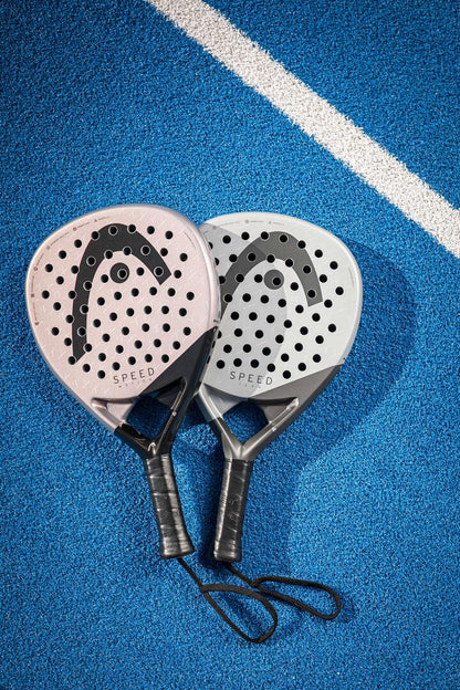 Two HEAD Speed Team 2025 Padel Rackets lie side by side on a blue court, showcasing Auxetic 2.0 technology. One is pink, the other grey/black, both branded "SPEED." A white line runs diagonally in the background, enhancing the scene's dynamic energy. Experience superior play with HEAD.